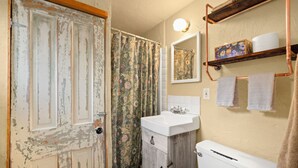 A bathroom with a rustic door, a shower with floral curtains, a white sink, a wooden cabinet, a white toilet, a mirror, and a towel rack with towels. - Estrella De Oro Residences: Casita- Walk to town! (Golden)