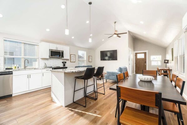 A modern kitchen with white cabinets, a central island, and bar stools. There is a dining area with a wooden table and chairs.