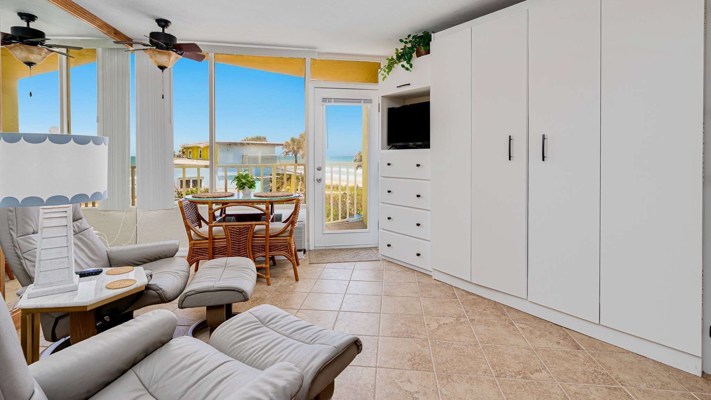 A modern living room with a sofa set, a dining table with chairs, a TV mounted on a cabinet, and a view of the sea through large windows.