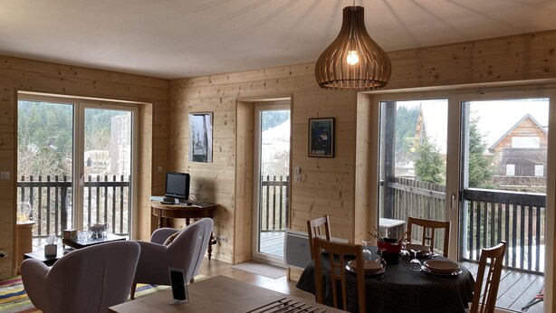 Una habitación acogedora con detalles de madera, una mesa de comedor con sillas, un ordenador sobre un escritorio y ventanas amplias que ofrecen vistas al exterior.