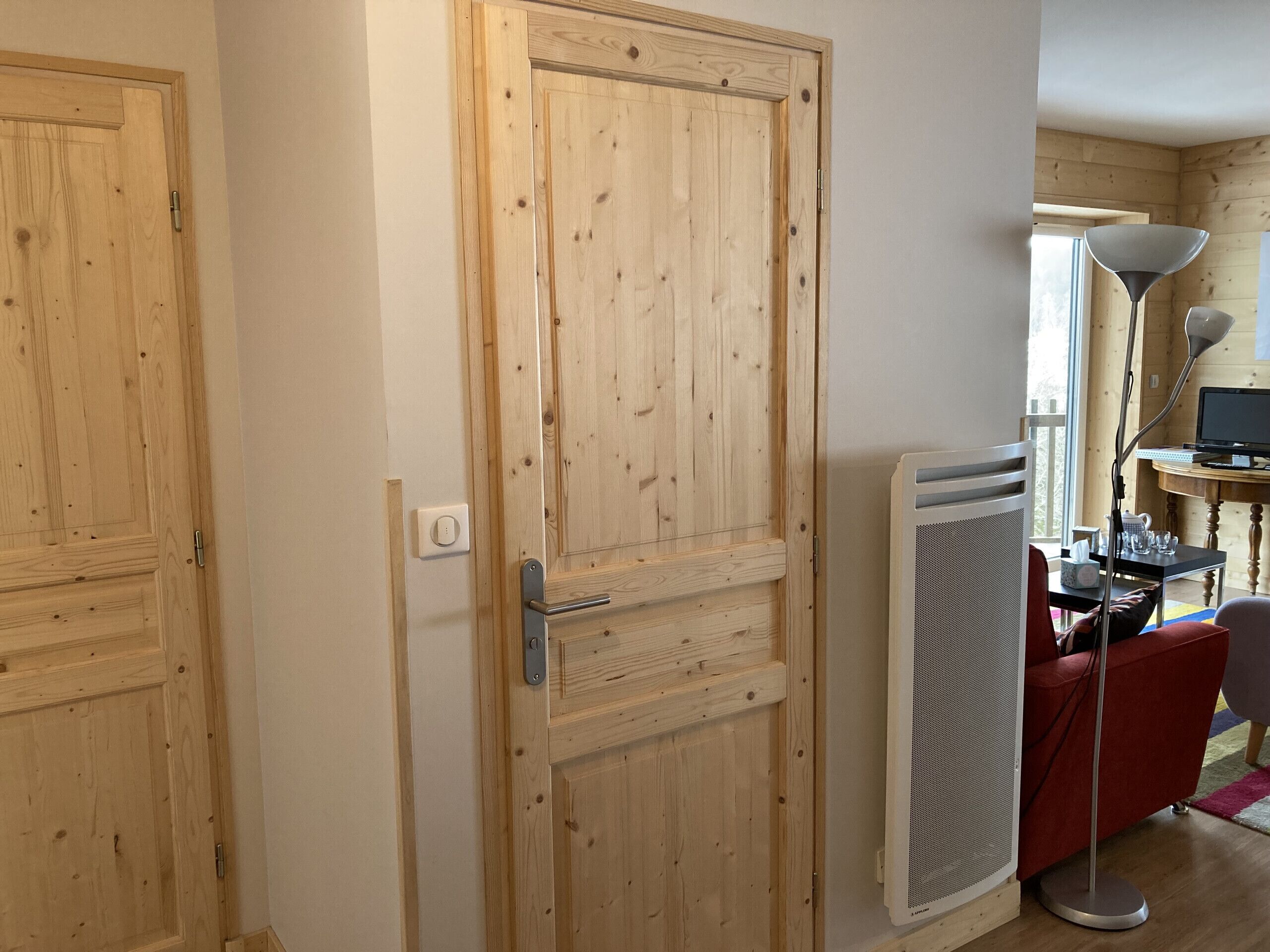 A wooden door with a handle and a light switch, a radiator, a desk with a computer, and a chair.