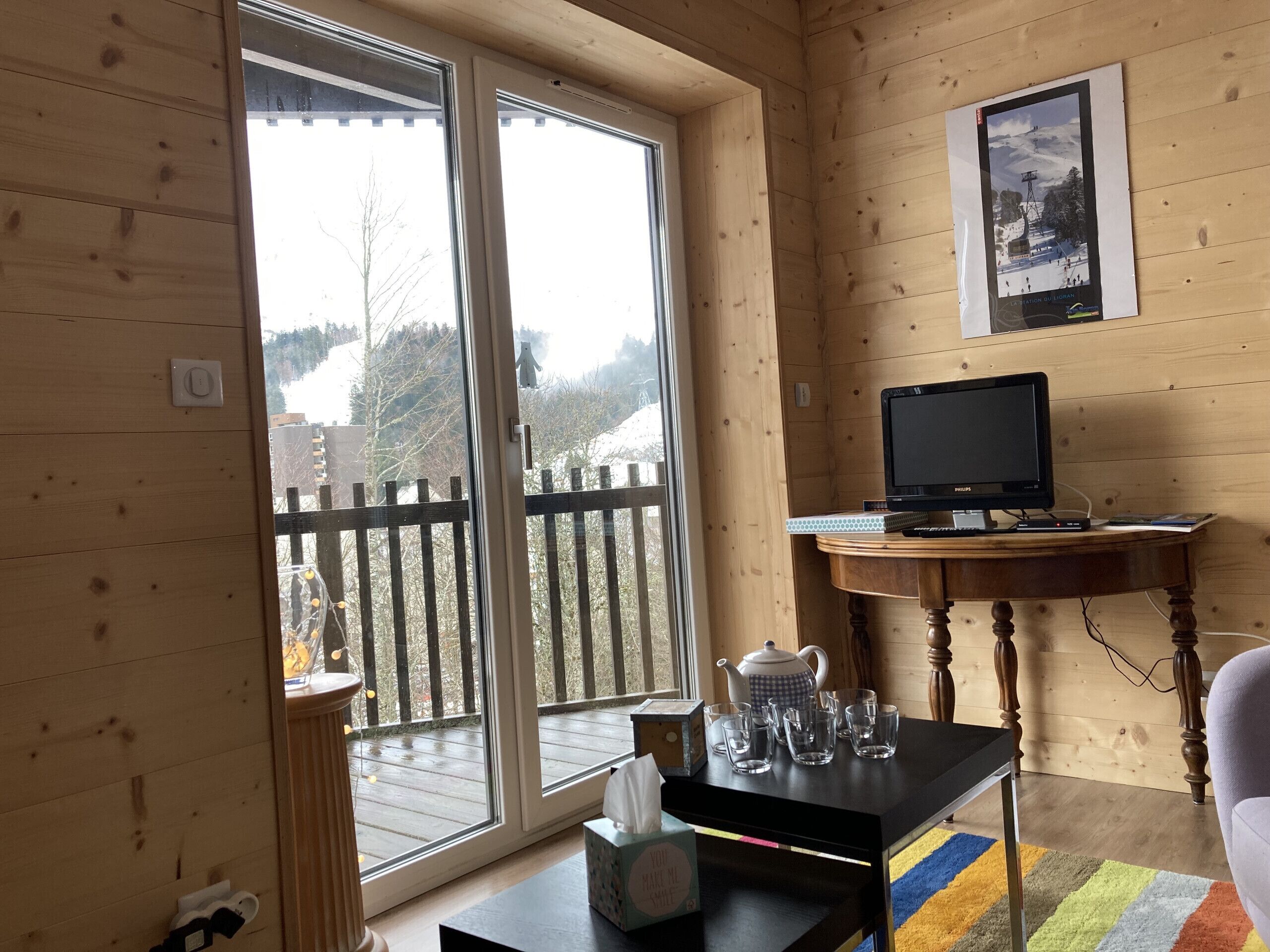 A cozy room with wooden walls, a large window with a view of a snowy landscape, a table with a computer, a teapot, and glasses.