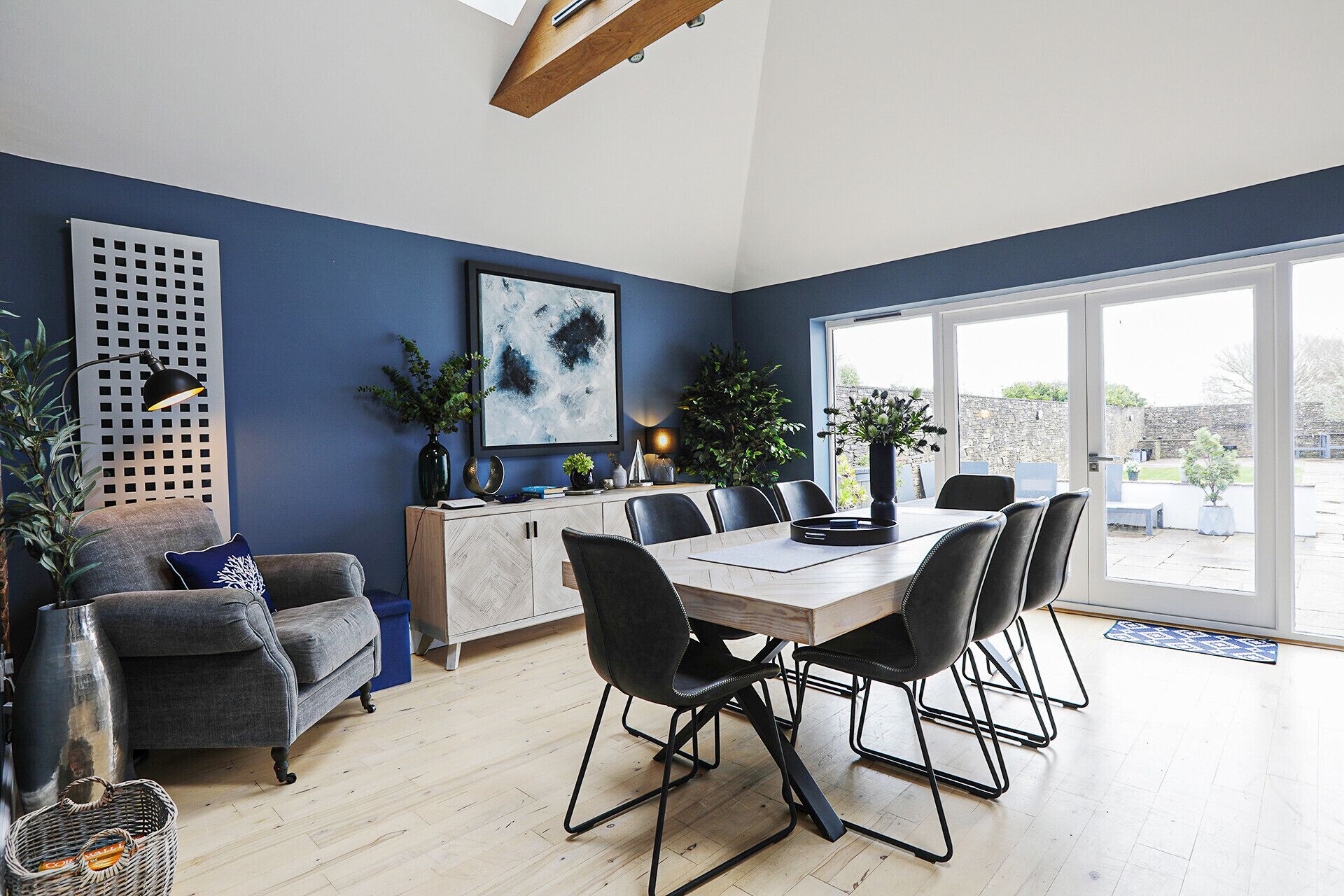 A modern dining area with a wooden table and black chairs, a blue sofa, and a large window offering a view of the outdoors.
