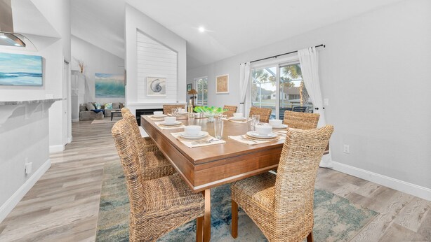 A dining area with a wooden table set for six, surrounded by wicker chairs, in a room with white walls and a wooden floor. - Canal-front Heated Pool Home with Pool Table & Kayaks (Palm Coast)