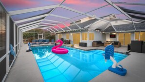 An indoor swimming pool with a glass ceiling, a pink flamingo float, and a blue diving board. - Canal-front Heated Pool Home with Pool Table & Kayaks (Palm Coast)
