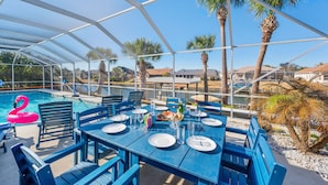 An outdoor dining area with blue tables and chairs, a pool with a pink flamingo float, and palm trees. - Canal-front Heated Pool Home with Pool Table & Kayaks (Palm Coast)