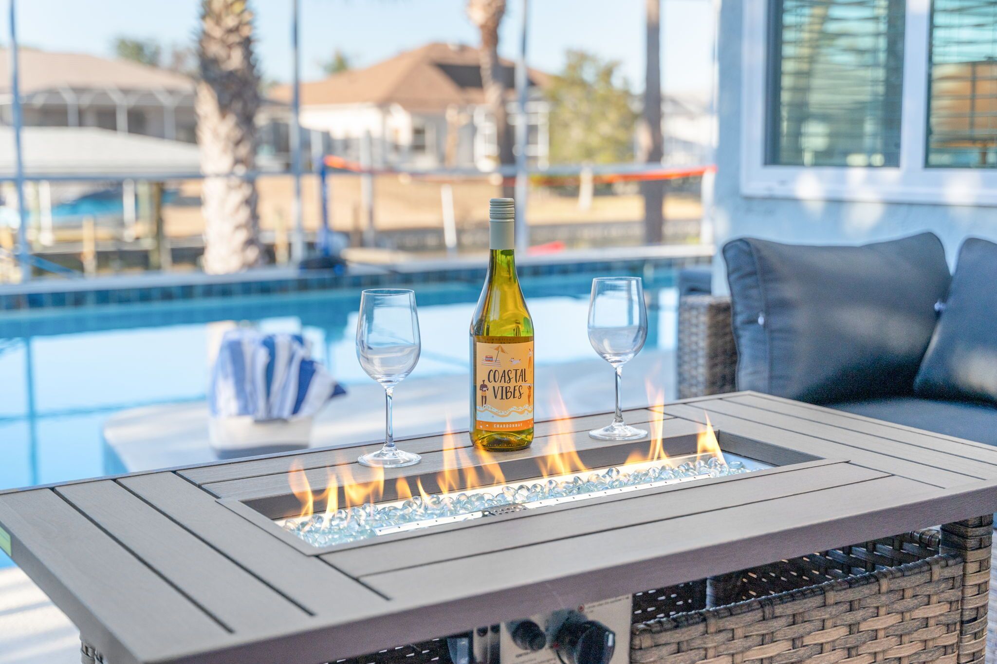 A poolside table with a bottle of Coastal Vibes wine, two glasses, and a fire feature.