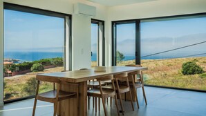 Un comedor con una mesa grande y sillas, ventanas amplias con vistas al mar y colinas.