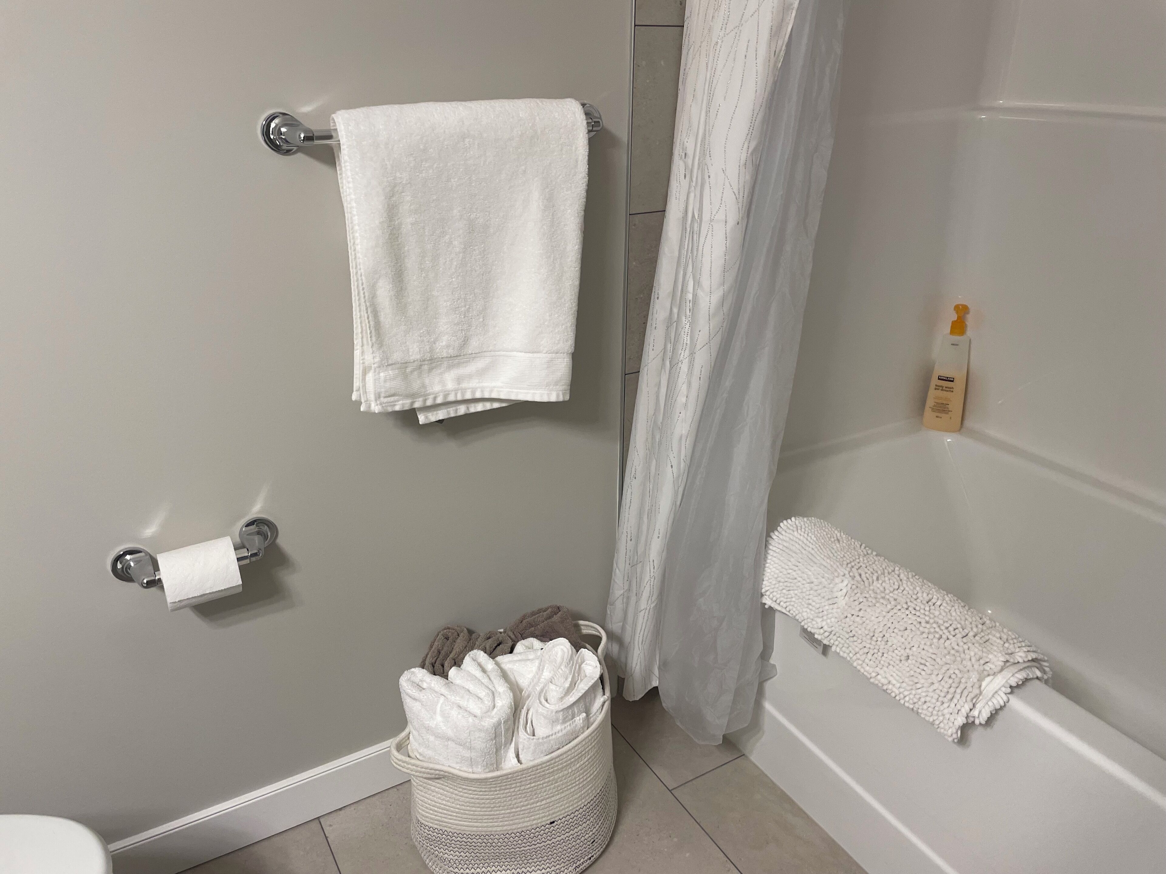 Ein Badezimmer mit einer weißen Badewanne, einer Duschvorhang, einem Handtuchhalter mit einem Handtuch, einem Korb mit gefalteten Handtüchern und einer Shampoo-Flasche.
