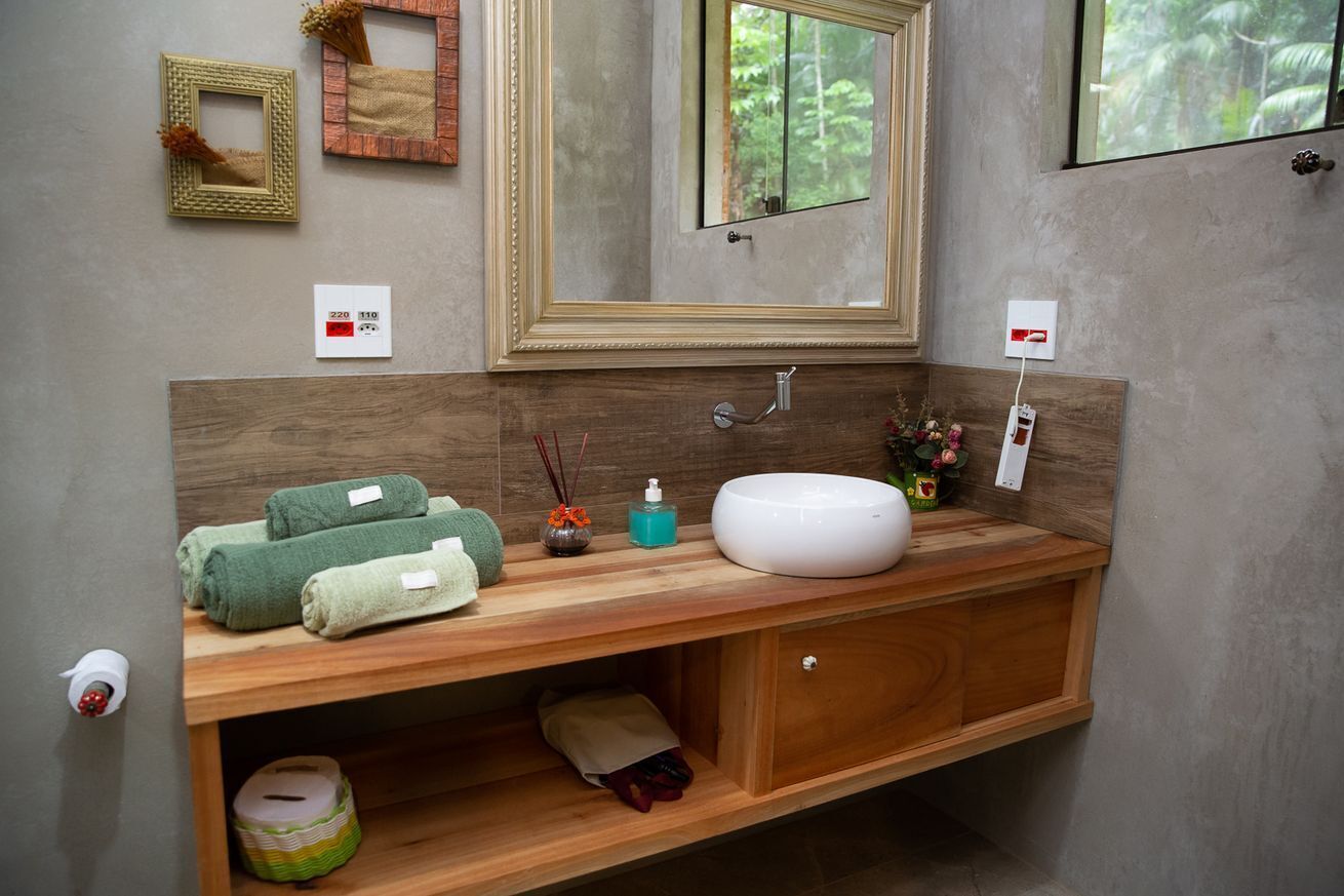 Une salle de bain avec un meuble-lavabo en bois, un lavabo blanc et un miroir. On y trouve des serviettes et un petit vase posé sur le meuble-lavabo.