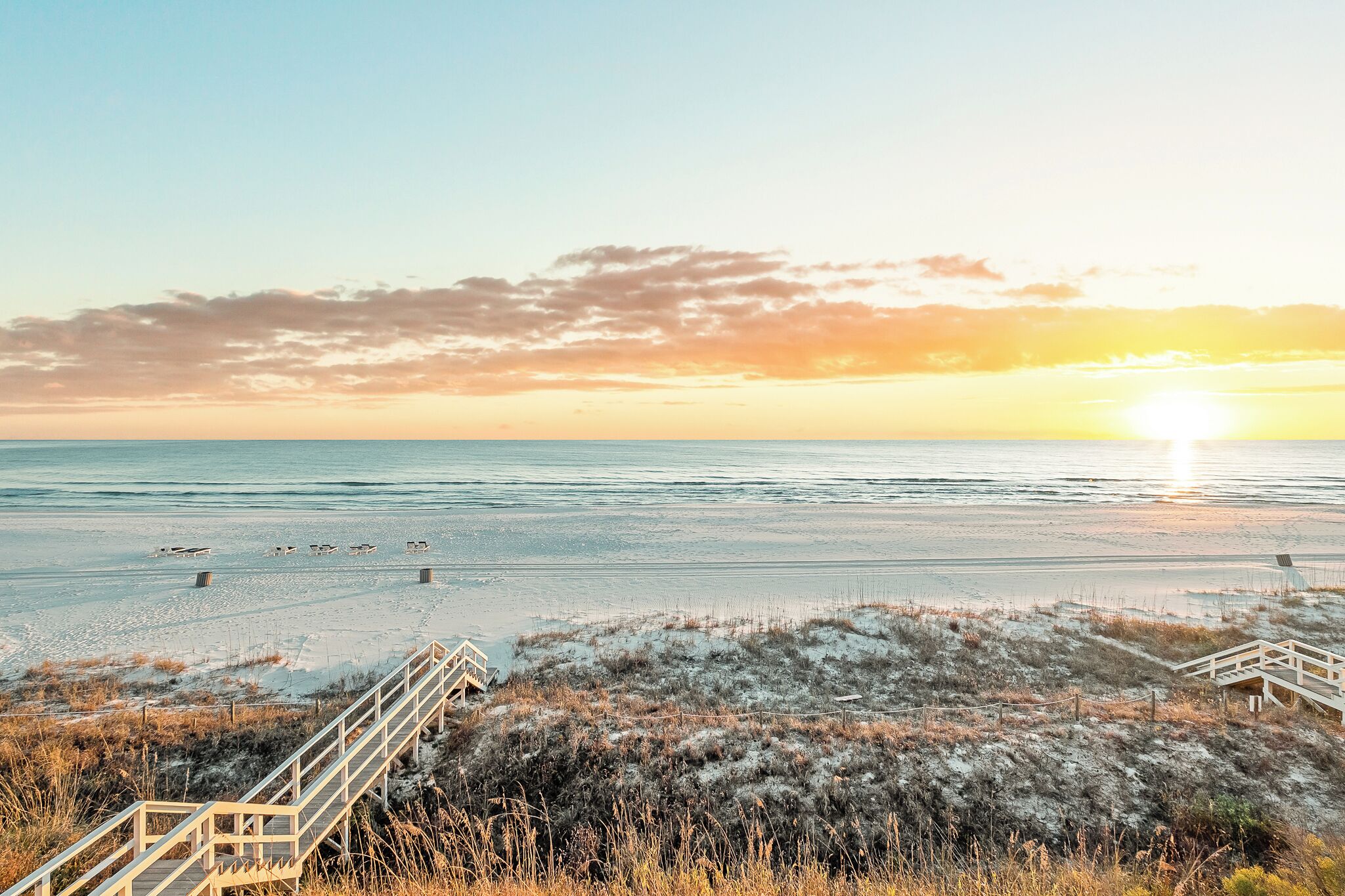 Ein Strand mit einer Holztreppe, die zum Wasser hinabführt, einem klaren Himmel und der untergehenden Sonne am Horizont.