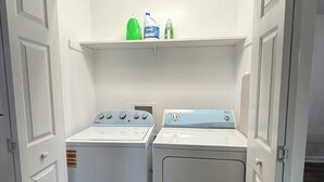 A laundry room with two front-loading washers and dryers, a shelf with cleaning supplies, and a white door. - Manning Way (Elon)