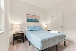 Un dormitorio con cama, dos mesitas de noche, una mesita auxiliar, una puerta y un cuadro colgado en la pared.