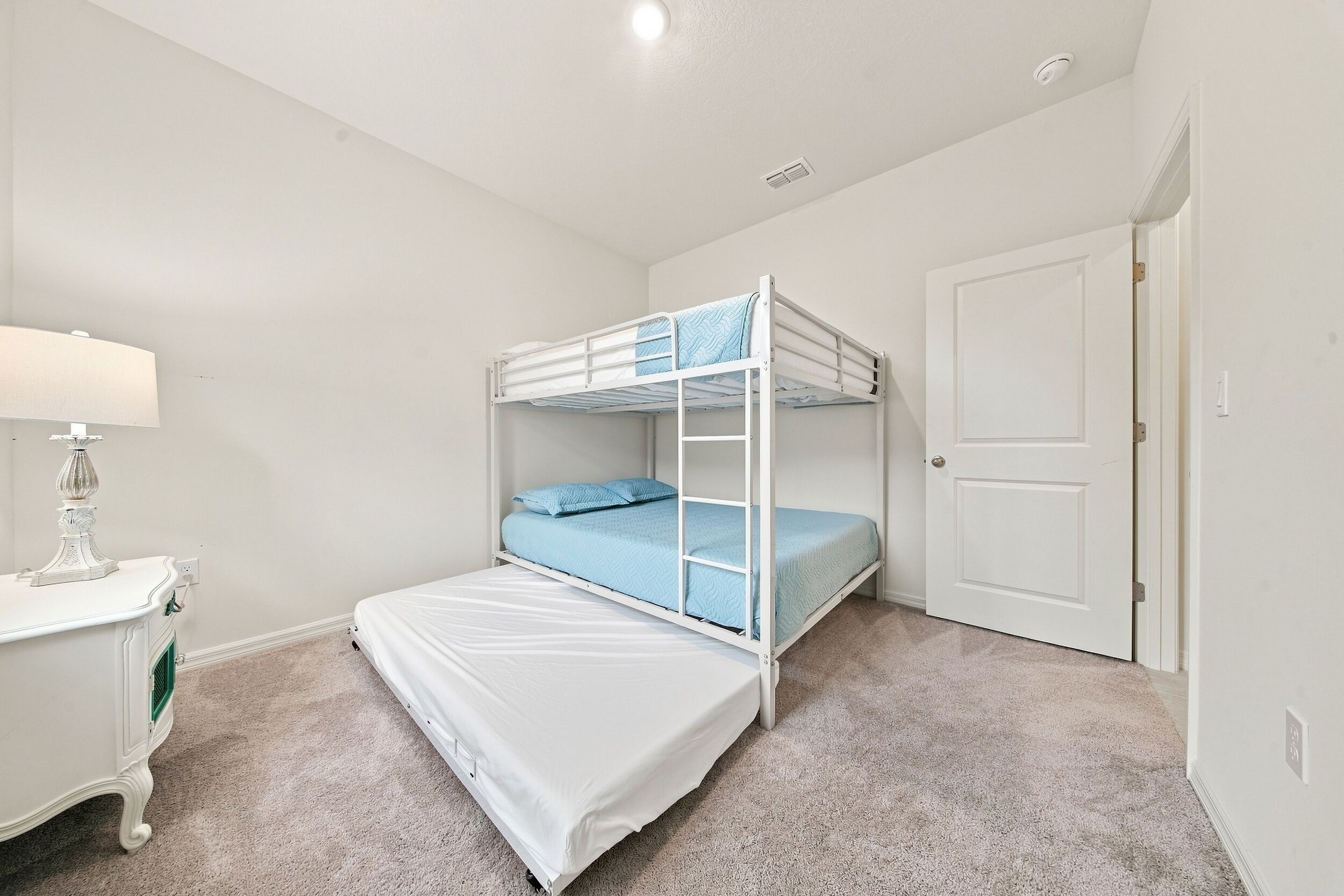 Ein Etagenbett mit weißem Gestell und blauer Bettwäsche, ein Nachttisch in Weiß mit einer Lampe und eine Tür im Zimmer.