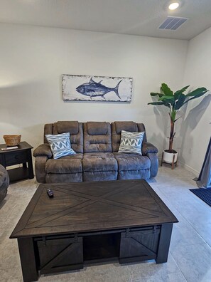A living room with a brown leather sofa, two patterned throw pillows, a wooden coffee table, a wall-mounted fish painting, a potted plant, and a small side table with a basket.