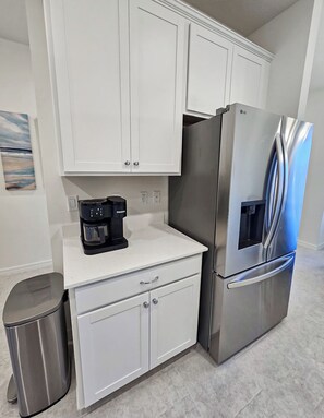 A modern kitchen with white cabinets, a stainless steel refrigerator, a coffee maker, and a trash can.