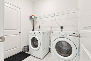 A laundry room with two front-loading washing machines, a drying rack, and a small trash bin.