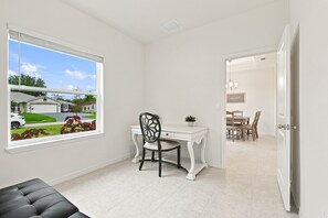 Una habitación con escritorio, una silla, un ventanal con vistas a una casa y una puerta que da a otra habitación.