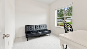 Quarto com sofá de couro preto, mesa branca e janela com vista para casas e árvores.
