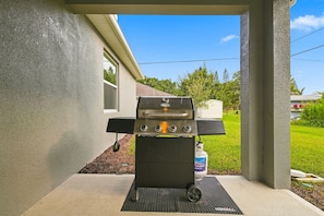 En bærbar gasgrill med fire brændere og en sidebrænder, placeret på en beton terrasse med en metaldrage.