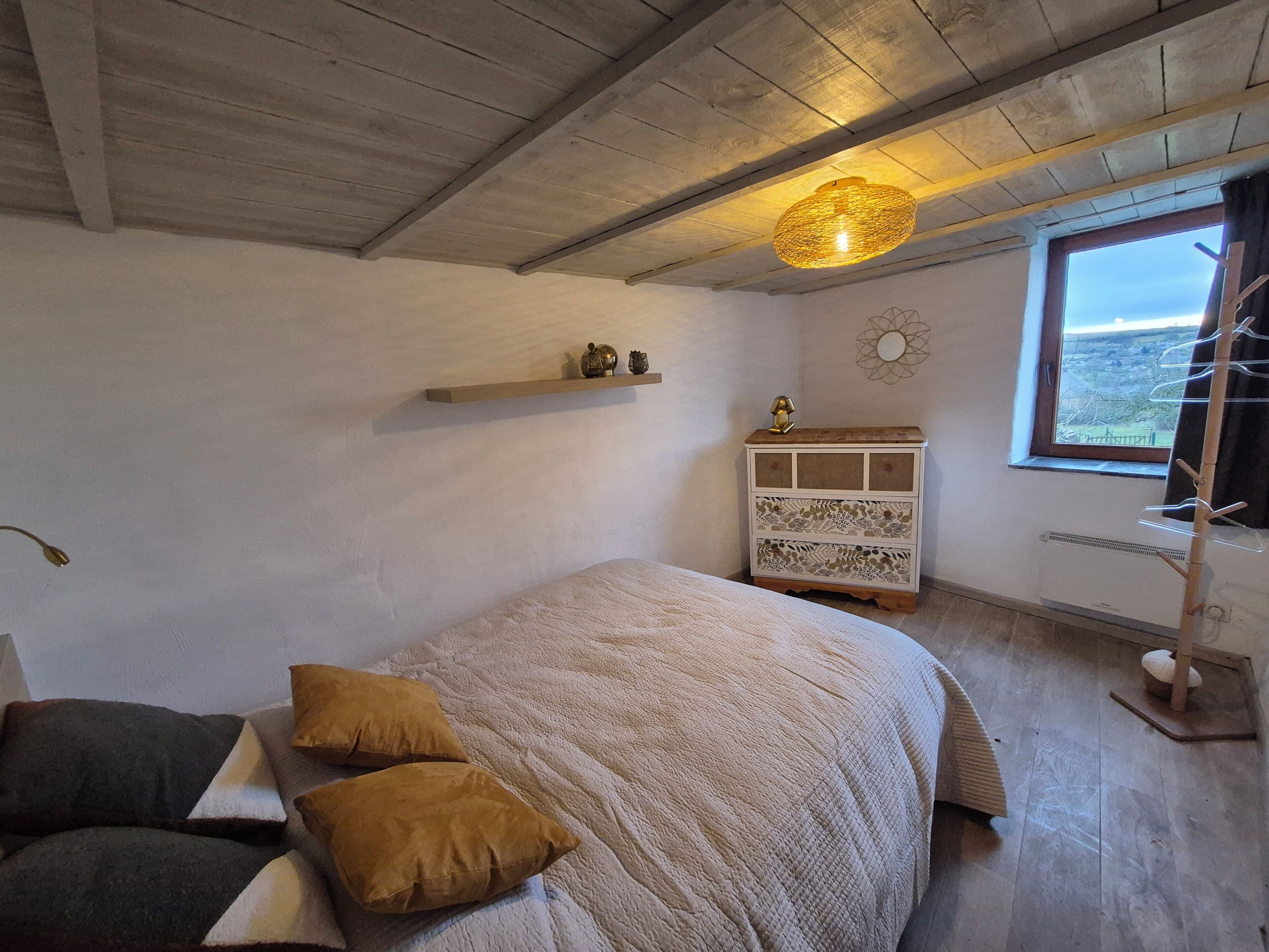 A bedroom with a bed, pillows, a nightstand, a wall-mounted shelf, a decorative wall piece, and a window with a view.