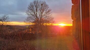 Solnedgang ses gennem et vindue med en stenvæg, et træ og en græsplæne.