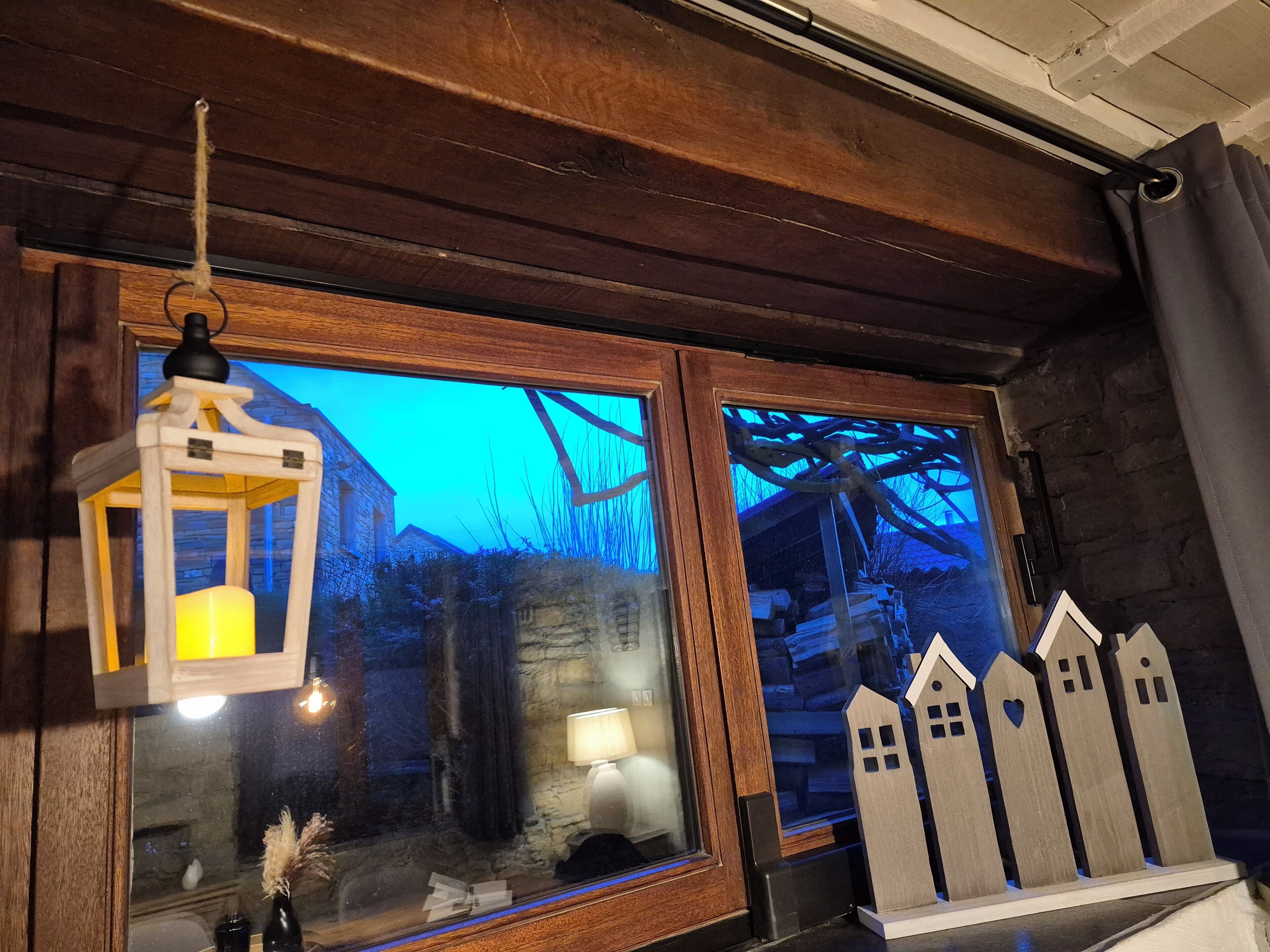 Une pièce rustique avec des poutres en bois, une lanterne, une fenêtre donnant sur un bâtiment en pierre et une petite table avec une lampe et des décorations représentant des maisons.