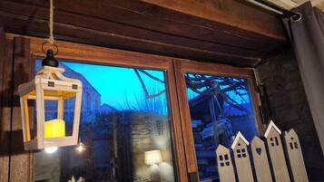 Une pièce rustique avec des poutres en bois, une lanterne, une fenêtre donnant sur un bâtiment en pierre et une petite table avec une lampe et des décorations représentant des maisons.