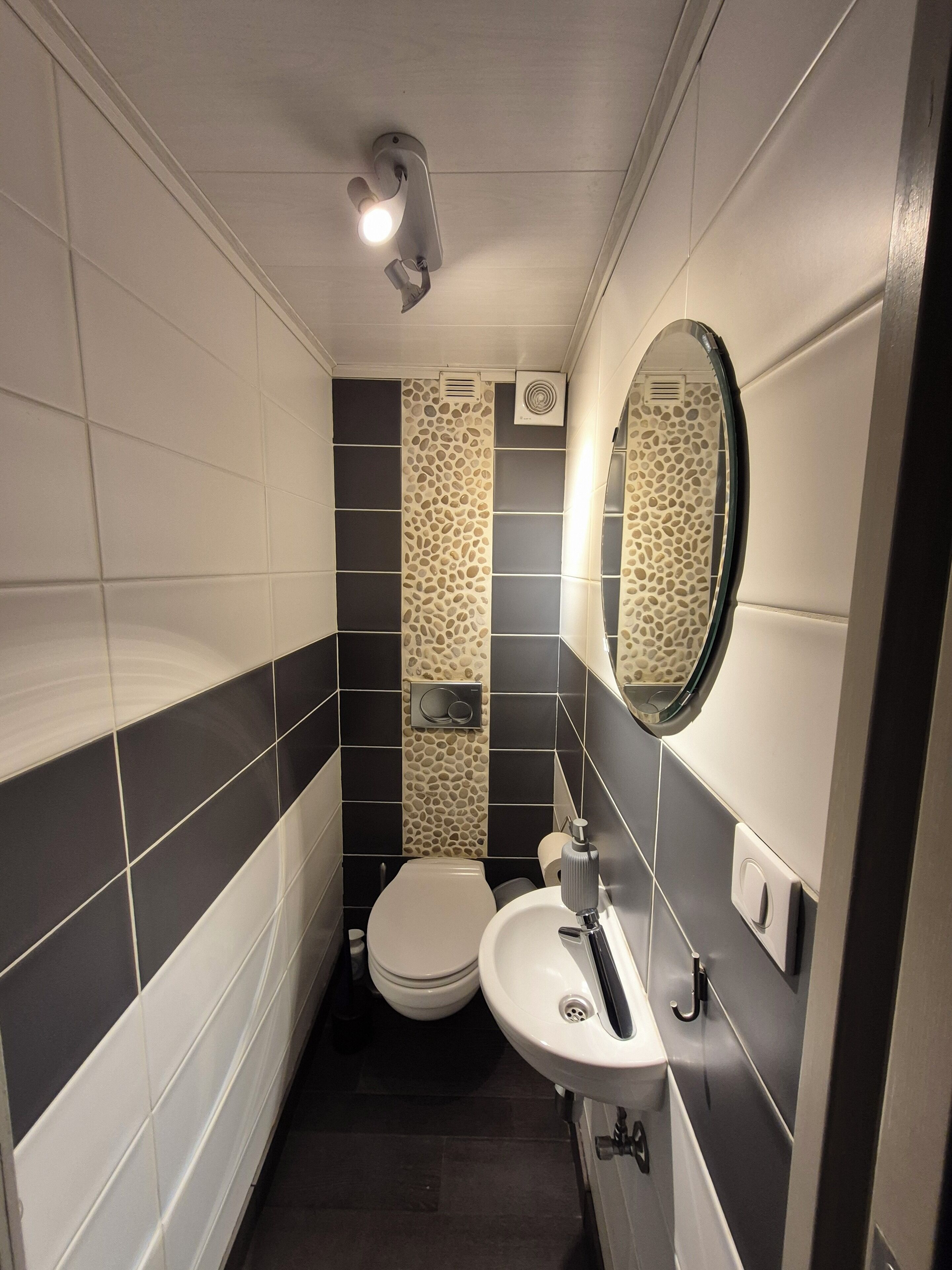 A compact bathroom with a toilet, sink, and mirror. It features a modern design with patterned tiles and a small ventilation unit on the wall.