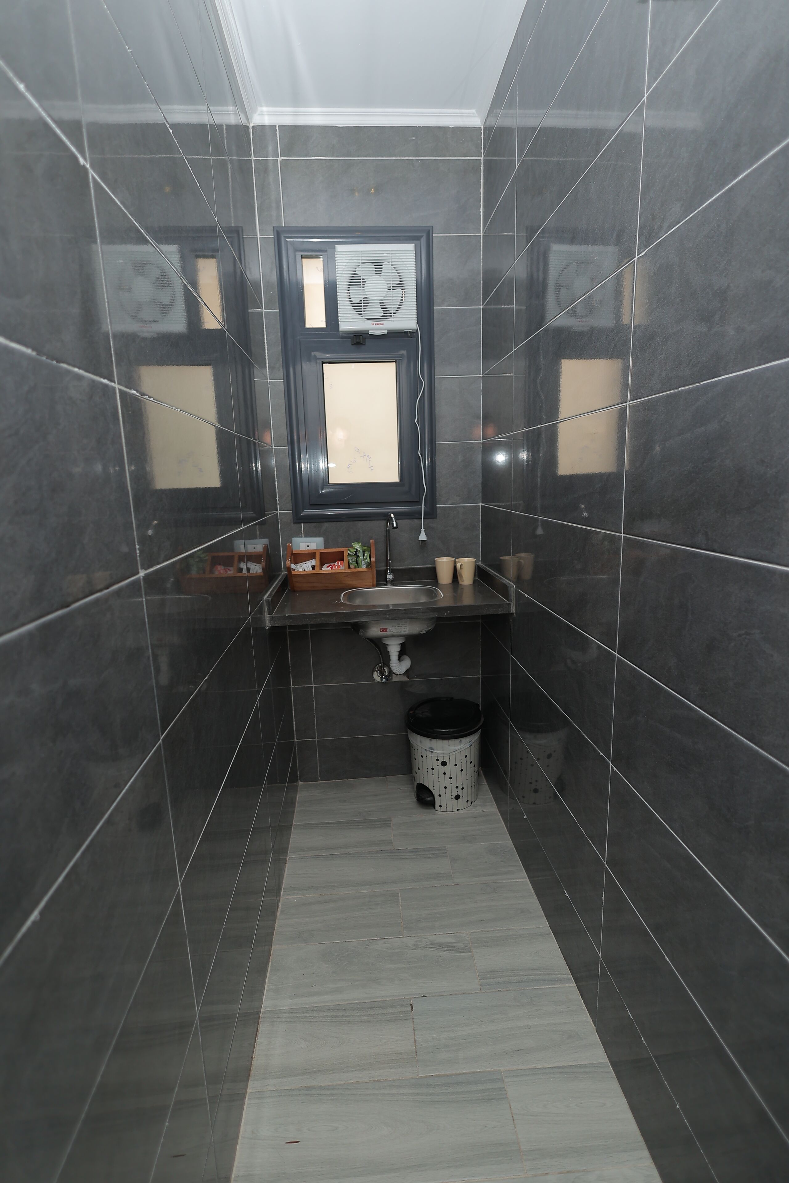 A narrow bathroom with dark tiles, a sink, a mirror, and a trash bin.