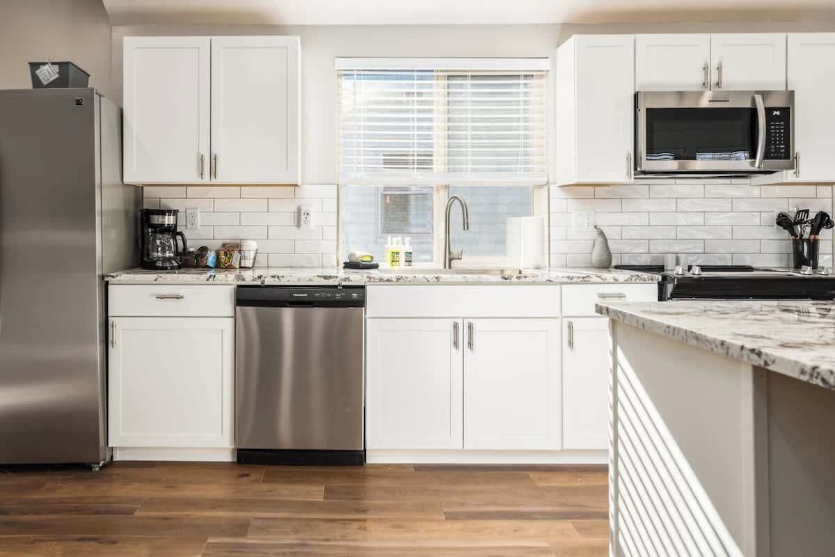 A modern kitchen with white cabinets, a stainless steel refrigerator, a dishwasher, a microwave, and a window with blinds.