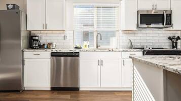 A modern kitchen with white cabinets, a stainless steel refrigerator, a dishwasher, a microwave, and a window with blinds.