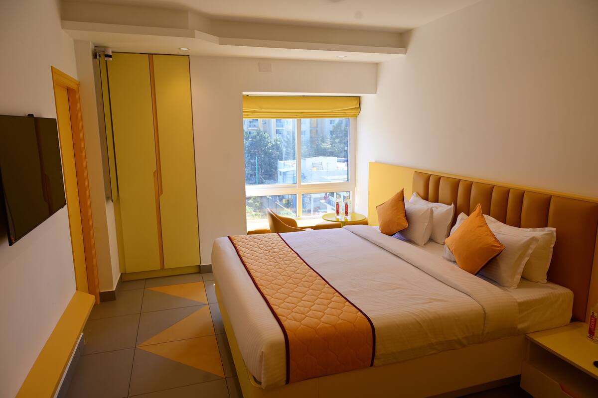 A hotel room with a large bed, a window with yellow curtains, a small table, and a TV mounted on the wall.