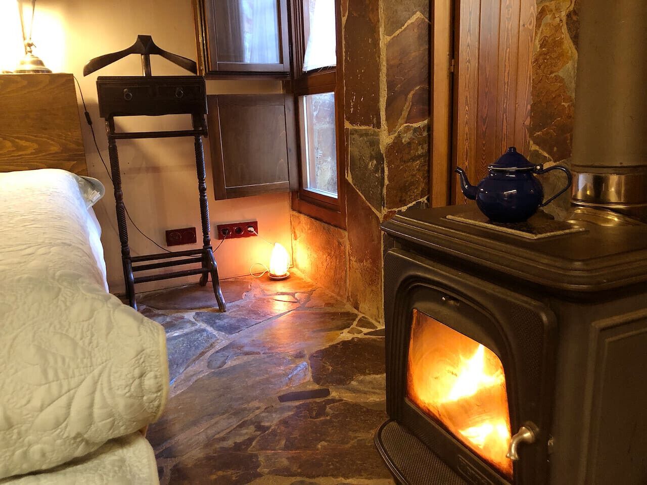 A cozy room with a stone floor, a wooden bed, a hanging chair, and a wood-burning stove.