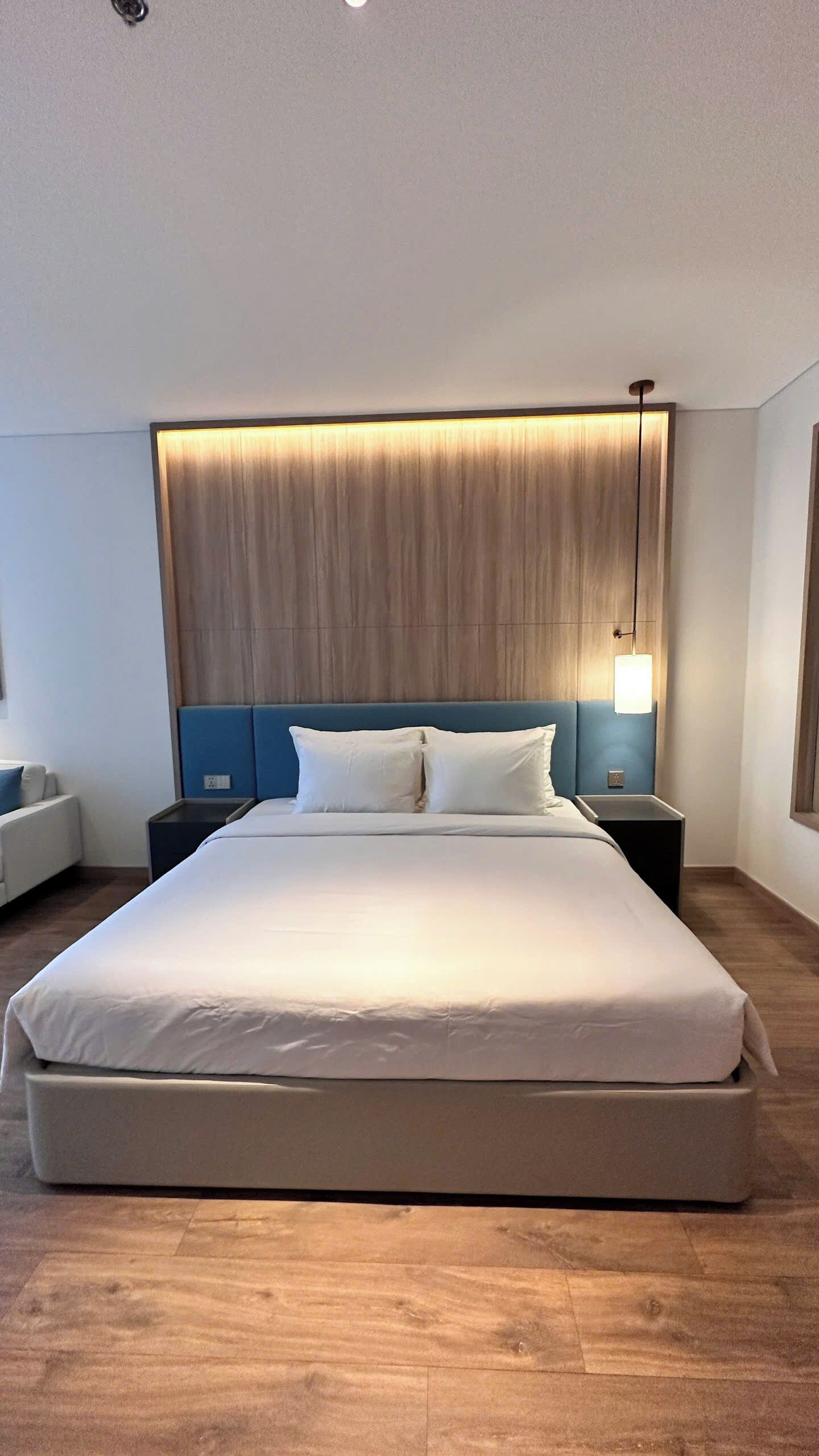 A modern hotel room with a large bed, wooden headboard, bedside tables, and a wall-mounted light fixture.