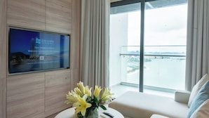 A modern hotel room with a large TV on the wall, a balcony with a view, and a vase of flowers on a round table. - A La carte Ha Long Apartment Sea View  (Bai Chay)