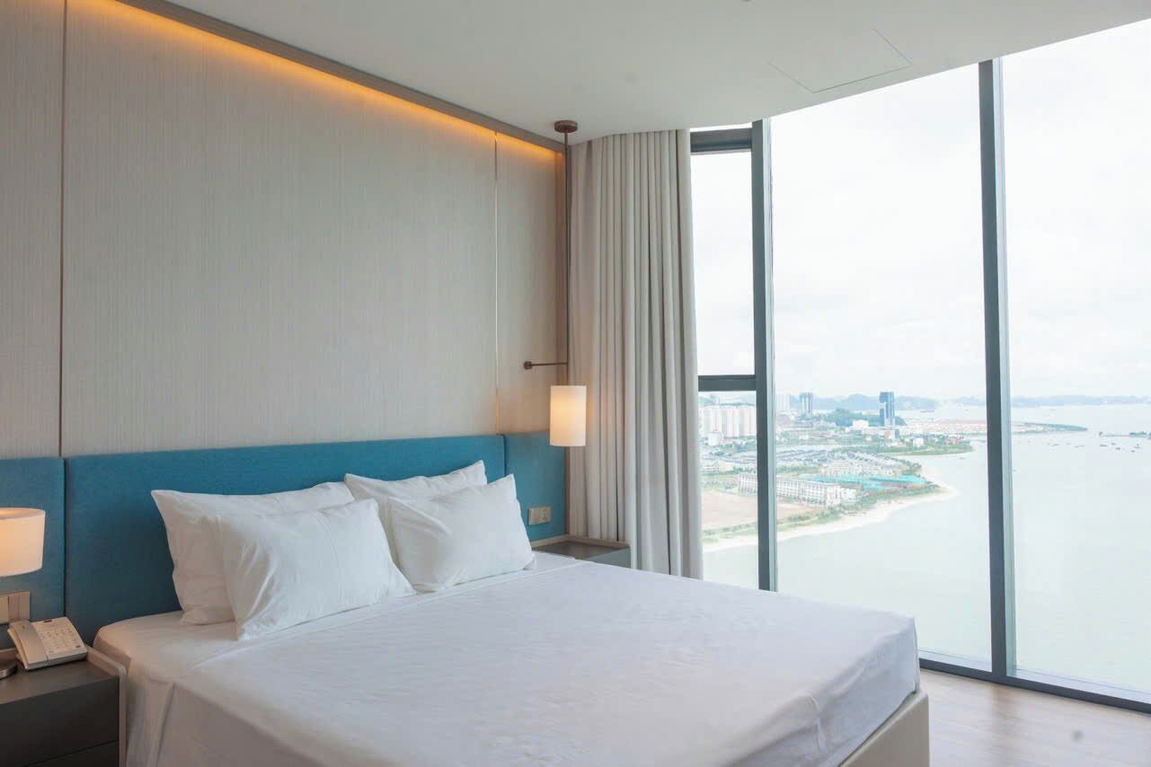 Ein Hotelzimmer mit einem großen Bett, einem Nachttisch mit Telefon, einem Fenster mit Blick auf Stadt und Wasser sowie einem blauen Kopfteil.