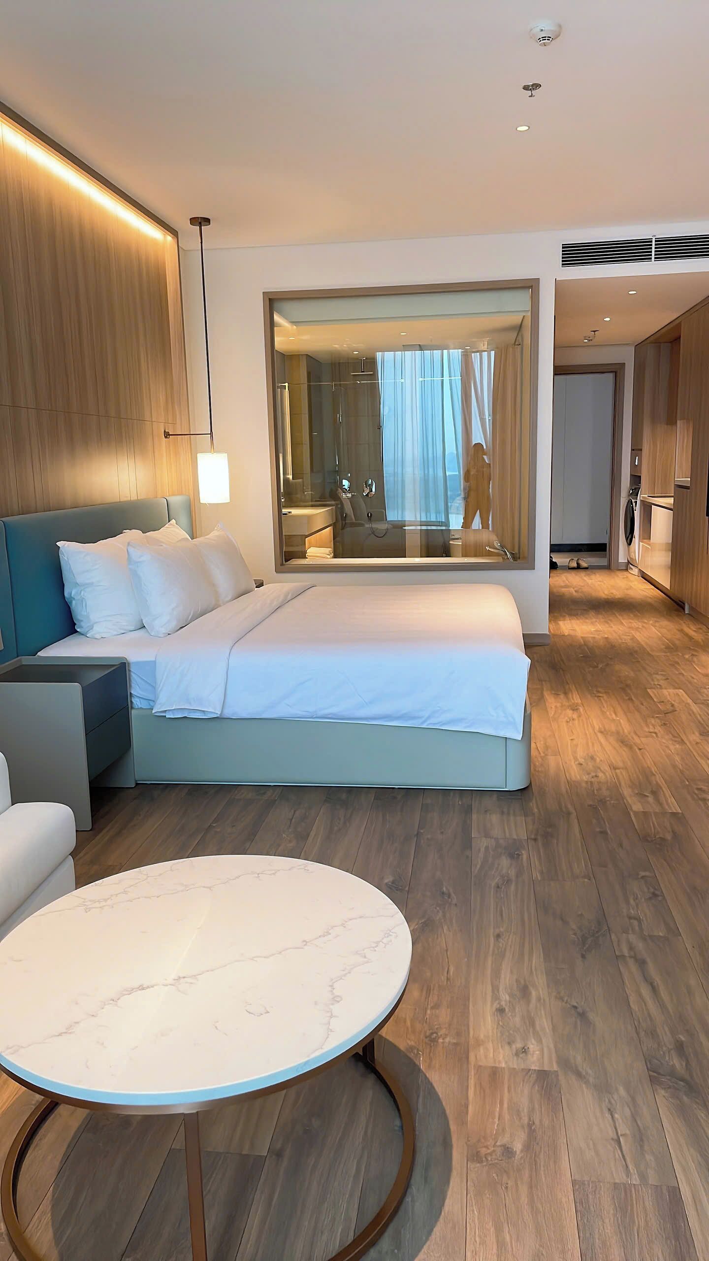 A modern hotel room with a large bed, a sitting area with a sofa, a round marble table, and a view of the bathroom through a large mirror.