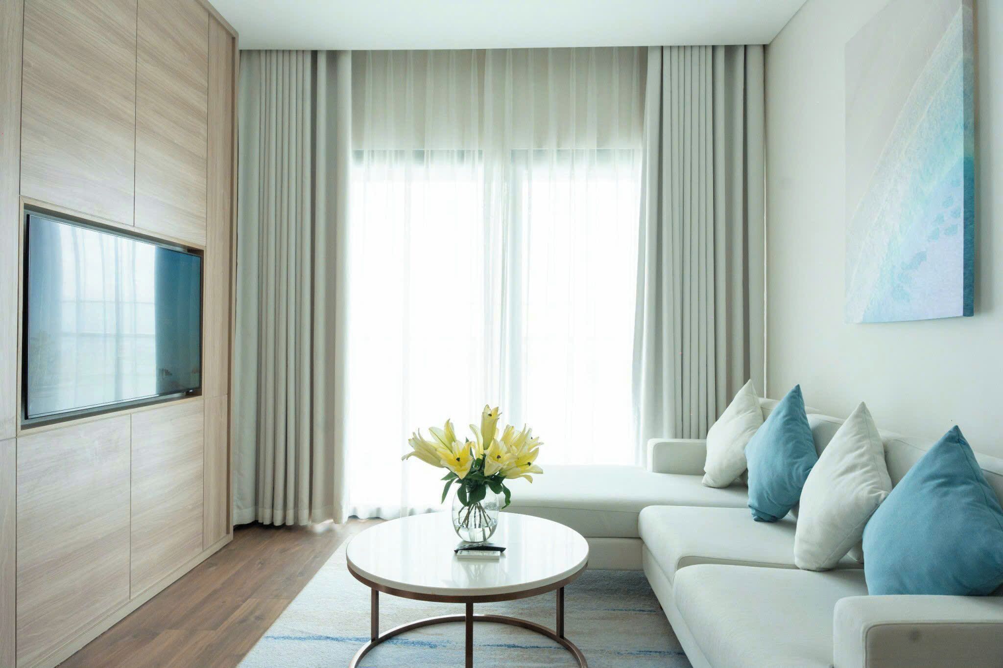 A modern living room with a flat-screen TV mounted on a wooden wall, a round coffee table with a vase of yellow flowers, a white sofa with blue cushions, and a large window with sheer curtains.