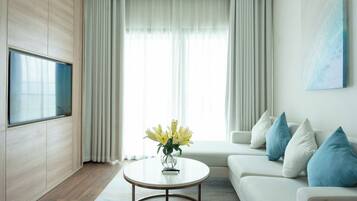 A modern living room with a flat-screen TV mounted on a wooden wall, a round coffee table with a vase of yellow flowers, a white sofa with blue cushions, and a large window with sheer curtains.
