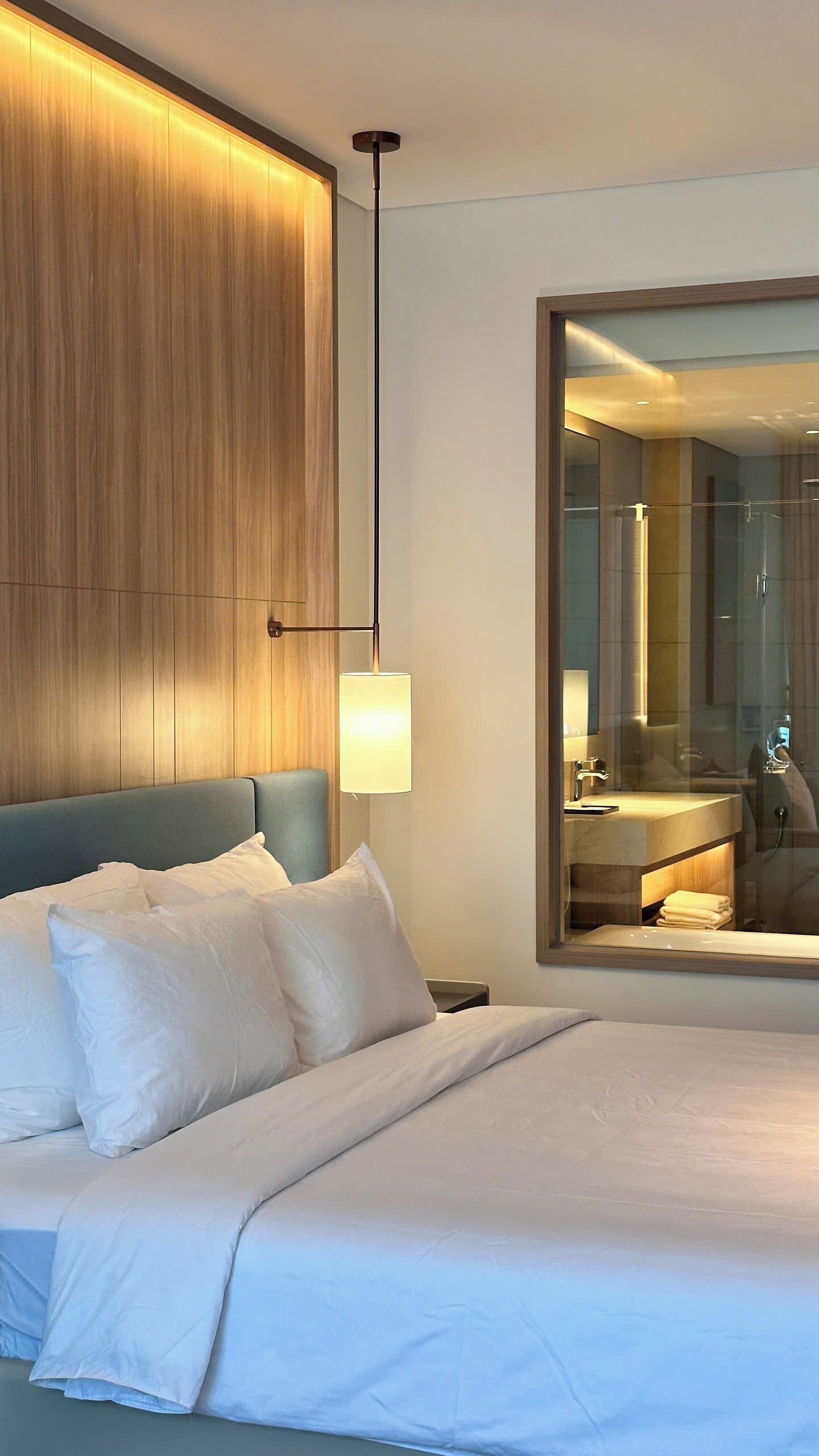 A modern hotel room with a large bed, a wooden panel wall, a large mirror, and a bedside lamp.