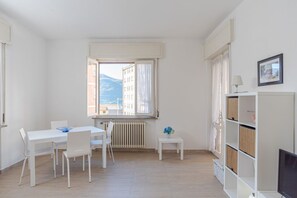 Una habitación con una mesa de comedor, sillas, una mesita auxiliar con un florero, un radiador, una ventana con cortinas, una estantería y un cuadro en la pared.