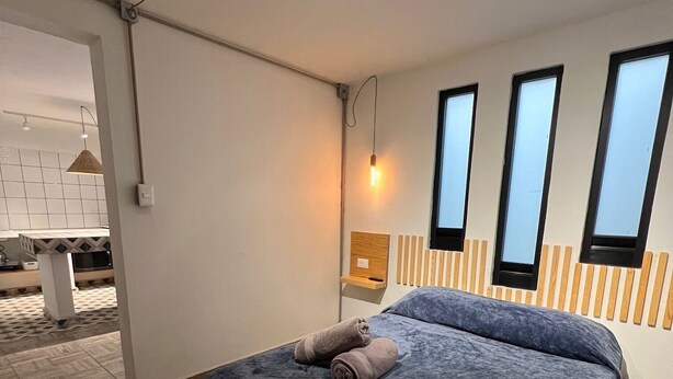 Et moderne soveværelse med en stor seng, en træsenggavl og et lille natbord. Der er tre vinduer i væggen, og en hængende lampe over sengen.