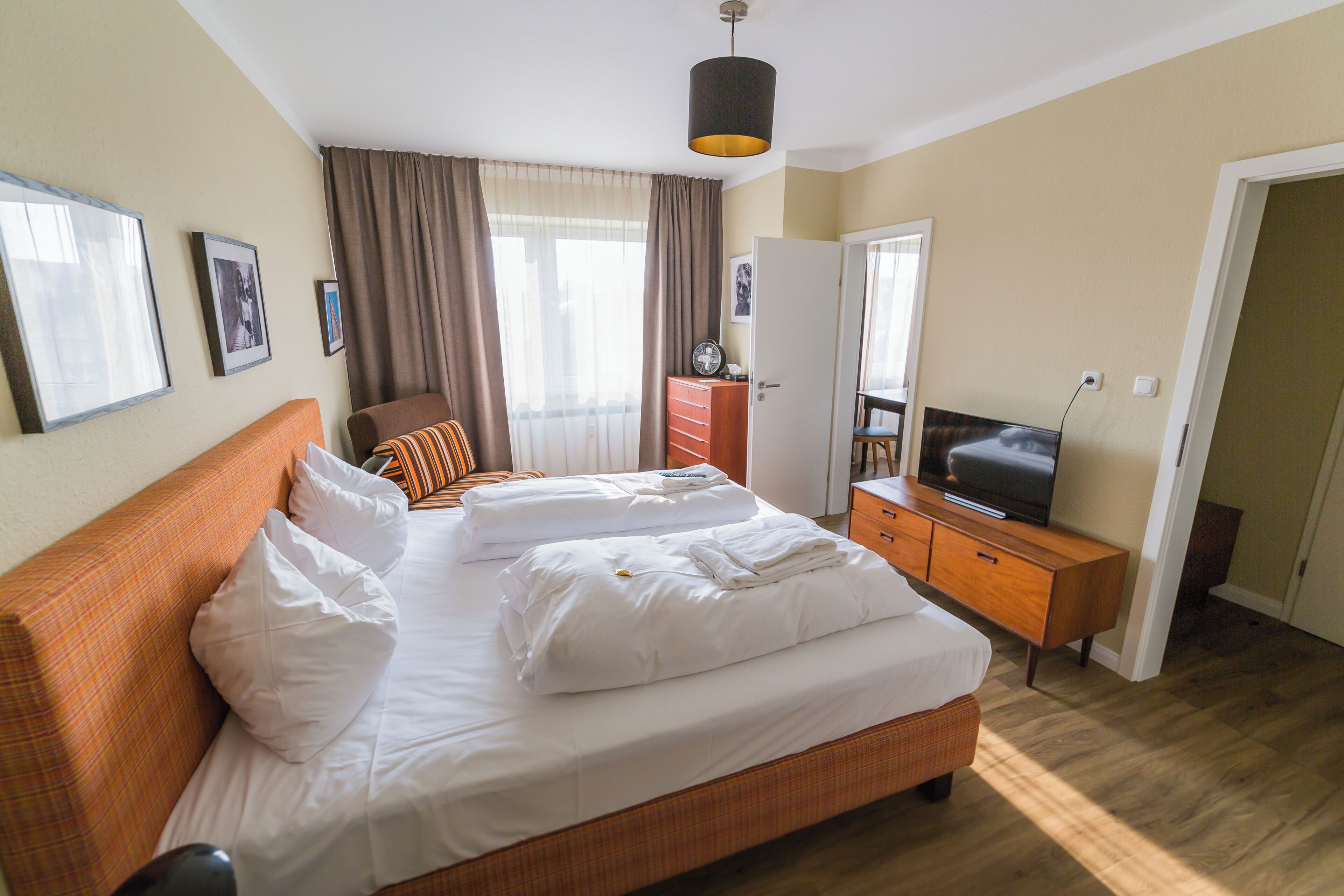 Una cama bien hecha con sábanas y almohadas blancas, un aparador de madera con un televisor y una puerta que da a otra habitación.