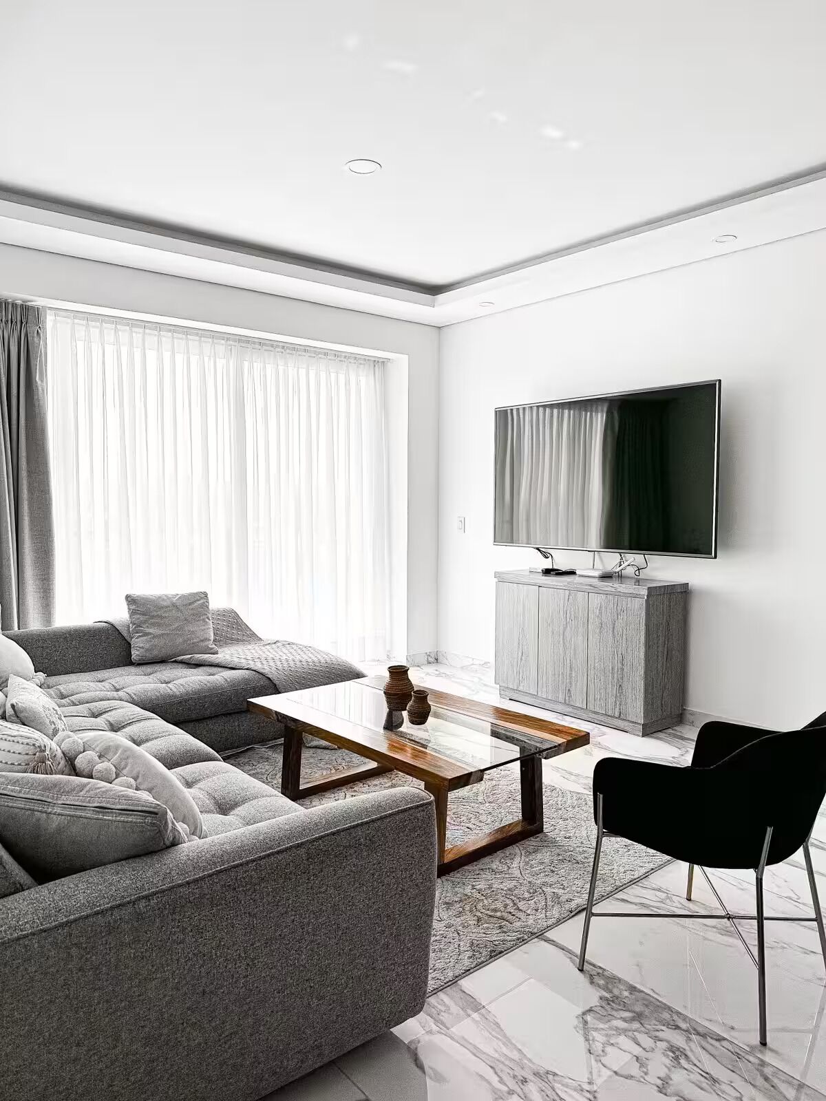 Een moderne woonkamer met een grijze modulaire bank, een houten salontafel, een zwarte fauteuil, een grijze kast en een flatscreen televisie die aan de muur is bevestigd.