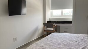 Ett sovrum med en säng, en tv som är monterad på väggen, ett fönster med persienner, en stol och en garderob.