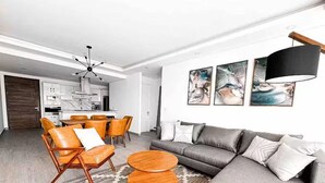 Een moderne woonkamer met een grijze bank, een houten salontafel en oranje leren fauteuils. Achtergrond is een keuken met witte kasten en een eethoek met een houten tafel en stoelen.