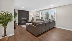 A modern living room with a grey sofa, a fireplace, and a large window. - Beaverton Escape, Backyard & Game Room (Beaverton)