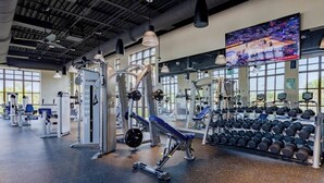 Una palestra con diverse attrezzature per esercizi, una televisione che mostra un evento sportivo e un espositore con manubri.
