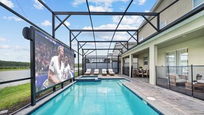 A swimming pool with lounge chairs, a covered seating area, and a large screen displaying a sports event. - 7BR house sleeps 20 with hot tub and pool (Kissimmee)